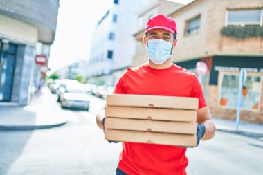 Üniforma giyen ve koronavirüs koruma maskesi takan genç teslimatçı. Kasaba caddesinde pizza kutularını teslim ediyorum..