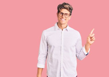 İş kıyafetleri giymiş, gözlüklü, yüzünde kocaman bir gülümseme olan genç ve yakışıklı bir adam kameraya el ile işaret ediyor.. 