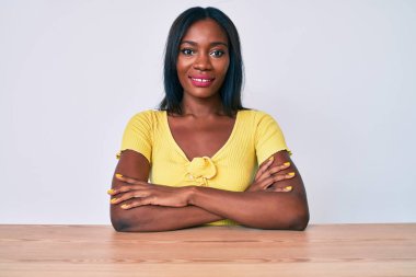 Genç Afro-Amerikan kadın gündelik giysiler içinde masaya oturmuş kollarını kavuşturup kameraya bakıyordu. pozitif kişi. 