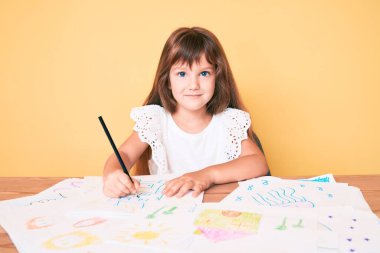 Uzun saçlı beyaz kız renkli resimler çiziyor. Pozitif ve mutlu görünüyor. Kendine güvenen bir gülümsemeyle dişlerini gösteriyor. 