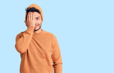 Yakışıklı Latin Amerikalı genç adam kışlık kazak giyiyor ve yün şapka takıyor. Bir gözünü eliyle kapatıyor. Yüzünde kendine güvenen bir gülümseme ve şaşırtıcı bir duygu var.. 