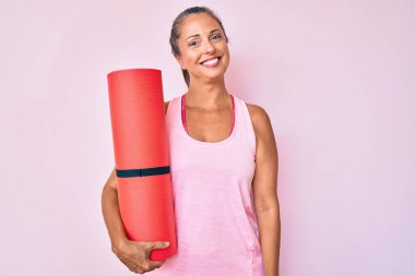 Orta yaşlı İspanyol kadın yoga minderi tutuyor. Olumlu ve mutlu görünüyor. Kendine güvenen bir gülümsemeyle dişlerini gösteriyor. 
