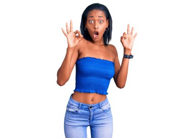 Genç Afrikalı Amerikalı kadın, günlük kıyafetlerini giymiş. Şaşırmış ve şok olmuş bir şekilde parmaklarıyla onay sembolü çiziyor. çılgın ifade 