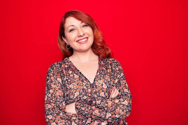 Kırmızı arka planda dikilip kameraya bakarken kollarını çapraz yapıp gülümseyen çiçekli bir elbise giyen güzel, kızıl saçlı bir kadın. Pozitif kişi.