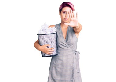 Pembe saçlı genç bir kadın. Elinde buruşuk kağıtlarla dolu bir çöp tenekesi var. Elinde dur işareti var. 