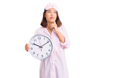 Uyku maskesi ve pijama giyen genç güzel Çinli kız büyük saati tutuyor ciddi bir yüz ifadesi takınıyor eli çenesinde soruyla ilgili sorular soruyor, kafa karıştırıcı bir fikir. 