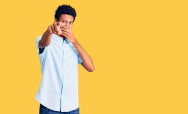 Gündelik giysiler giymiş genç Afro-Amerikalı adam sana gülüyor, eli ağzında kamerayı işaret ediyor, utanç ifadesi var. 