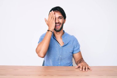 Yakışıklı İspanyol bir adam, gündelik giysiler içinde masada oturuyor bir gözünü eliyle kapatıyor, yüzünde kendine güvenen bir gülümseme ve sürpriz bir duygu var.. 