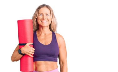 Orta yaşlı, sarışın, yoga minderi tutan, pozitif ve mutlu görünen, kendine güvenen, gülümseyen, dişlerini gösteren bir kadın. 