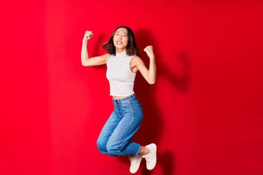 Genç, güzel Çinli kadın rahat kıyafetler giyip mutlu gülümsüyor. Yüzünde tebessümle zıplıyor, kırmızı arka planda yumruklarıyla kazanan jestler yapıyor.