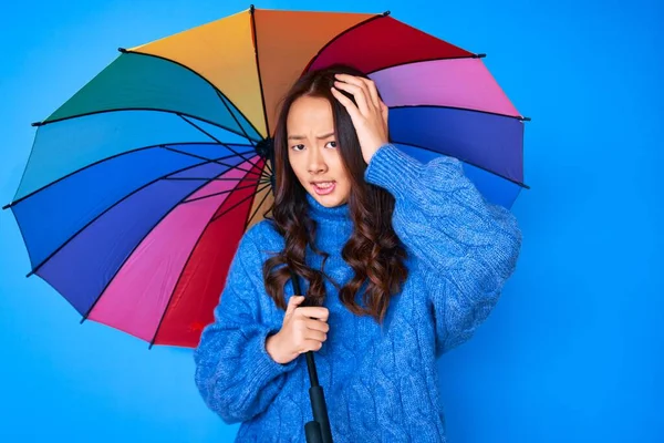 若いです美しいです中国の女の子保持カラフルな傘を強調し 頭の上に手で不満 驚きと怒っている顔 — ストック写真