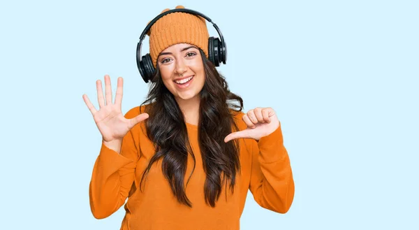 Beautiful brunette young woman listening to music using headphones showing and pointing up with fingers number six while smiling confident and happy.