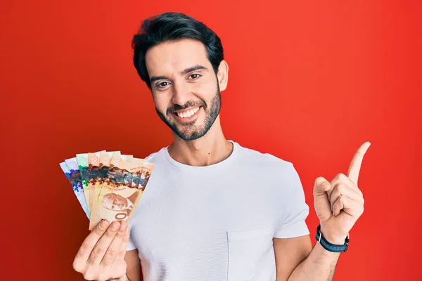 Ung Latinamerikan Man Håller Kanadensiska Dollar Ler Glad Pekar Med — Stockfoto