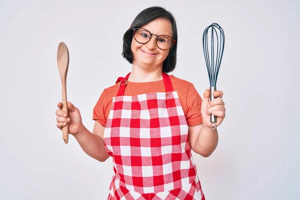 Brunette woman with down syndrome cooking using baker whisk and spoon smiling and laughing hard out loud because funny crazy joke.