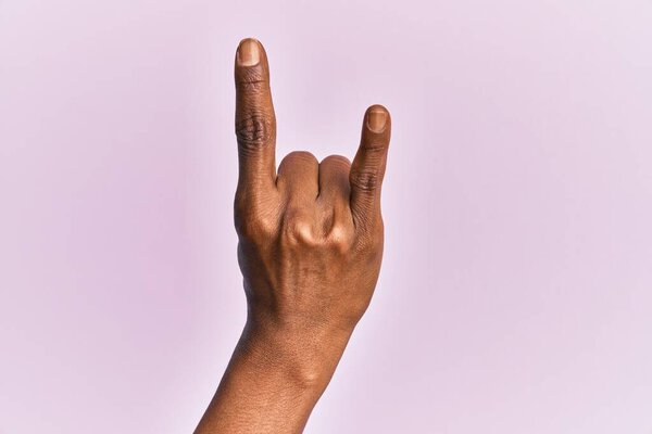 Arm and hand of black middle age woman over pink isolated background gesturing rock and roll symbol, showing obscene horns gesture 