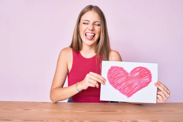 Joven Chica Rubia Sentada Mesa Sosteniendo Corazón Dibujar Lengua Que —  Fotos de Stock