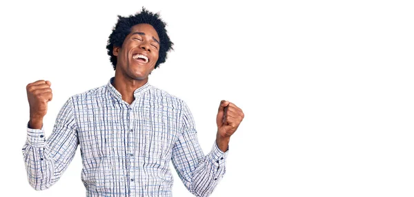 アフロの髪をしたハンサムなアフリカ系アメリカ人の男性は カジュアルな服を着て非常に満足し 腕を上げ 笑顔と成功のために叫んで勝者のジェスチャーを行う興奮しています お祝いのコンセプト — ストック写真