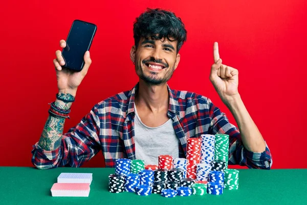 Молодий Іспаномовний Чоловік Сидить Столі Покерними Чіпсами Картами Тримають Смартфон — стокове фото
