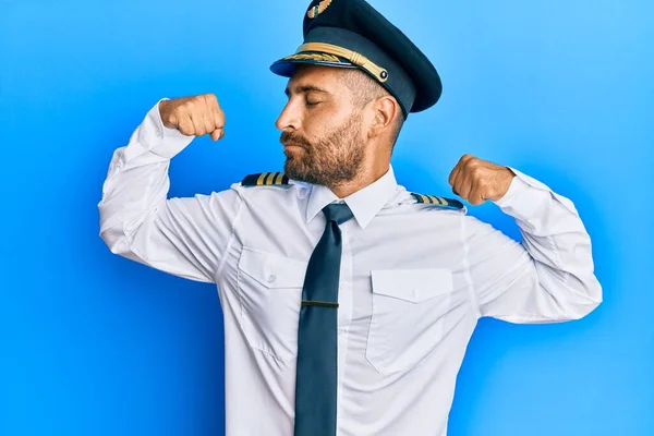 Handsome man with beard wearing airplane pilot uniform showing arms muscles smiling proud. fitness concept.