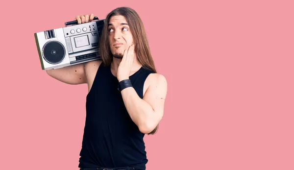 Young adult man with long hair wearing black clothes and holding boombox serious face thinking about question with hand on chin, thoughtful about confusing idea