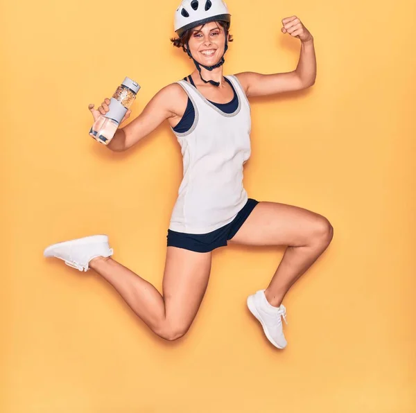 若い美しいバイカーの女性が幸せな笑顔自転車ヘルメットを着用 顔に笑顔でジャンプ隔離された黄色の背景に水のボトルを保持 — ストック写真