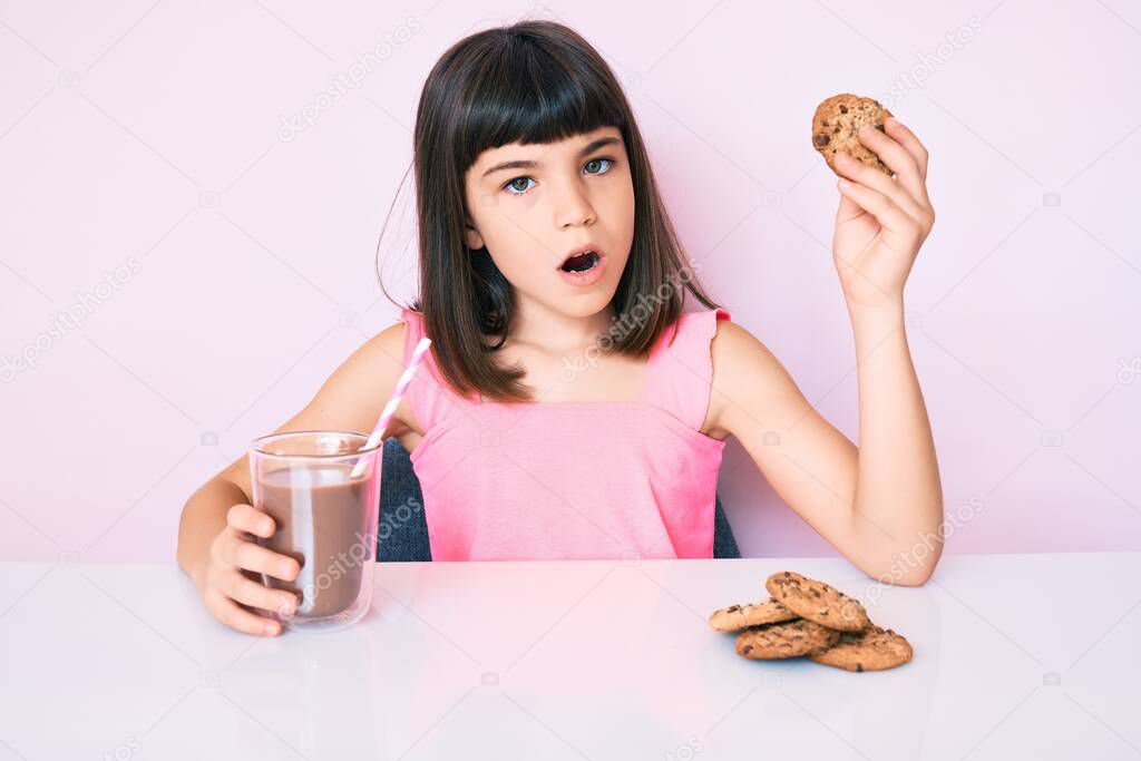 Young little girl with bang sitting on the table having breakfast in shock face, looking skeptical and sarcastic, surprised with open mouth 