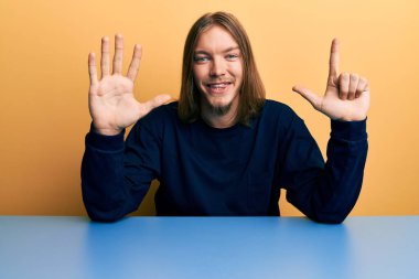 Uzun saçlı yakışıklı beyaz adam masadaki günlük kıyafetlerini giymiş yedi numaralı parmaklarıyla oturuyor ve gülümsüyor ve mutlu.. 