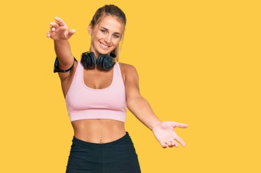 Spor kıyafetleri giyen ve kulaklık takan genç sarışın kadın kucaklanmak için kollarını açmış kameraya bakıyor. Mutluluğu kucaklayan neşeli bir ifade.. 