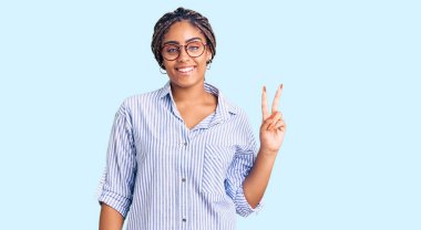 Saçları örgülü ve gözlüklü genç Afro-Amerikalı kadın iki numarayı gösterip gösterirken kendinden emin ve mutlu gülümsüyor.. 