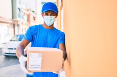 Kurye üniforması giyen Afrikalı kurye dışarıda. Koronavirüs güvenlik maskesi takıyor. Elinde karton bir paket var.