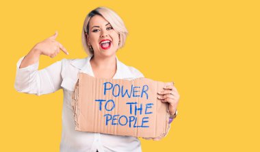 Genç sarışın ve cüsseli kadın gücü elinde tutuyor insanların afişlerini gösteriyor mutlu ve gururlu bir şekilde gülümseyen birini işaret ediyor. 