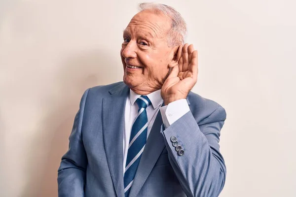 Senior handsome grey-haired businessman wearing elegant suit over white background smiling with hand over ear listening and hearing to rumor or gossip. Deafness concept.