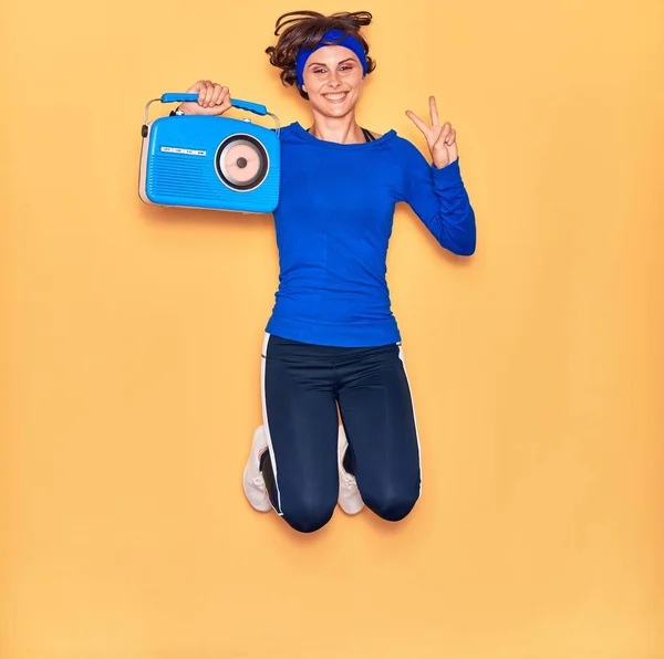 年轻美丽的女运动员开心地听着音乐 面带微笑地跳着 手持老式收音机 在孤立的黄色背景上做胜利的标志 — 图库照片