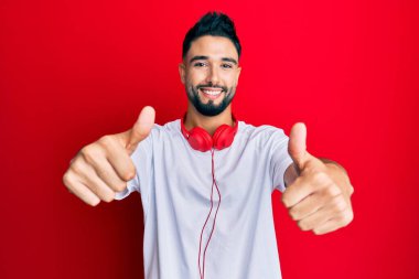 Sakallı genç adam kulaklık takarak müzik dinliyor. Elleriyle olumlu jest yapıyor. Gülümsüyor ve başarı için seviniyor. kazanan jest. 