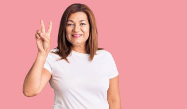 Orta yaşlı beyaz tişört giyen Latin kadın kameraya bakıp zafer işareti yaparken gülümsüyor. İki numara.. 