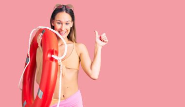 Güzel, esmer, bikini giyen, elinde can kurtaran yeleği tutan mutlu ve pozitif gülümseyen, başparmağını kaldıran, mükemmel ve onay işareti yapan genç bir kadın. 