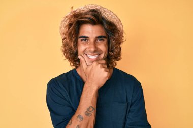 Yaz şapkası takan İspanyol genç adam kameraya kendinden emin bakıyor kollarını kavuşturup gülümsüyor ve çenesini kaldırıyor. Olumlu düşünmek. 