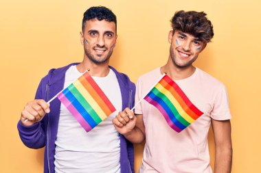 Genç eşcinsel çift ellerinde gökkuşağı lgbtq bayrakları tutarken pozitif ve mutlu görünüyorlar. Dişlerini gösteren bir gülümsemeyle ayakta duruyorlar. 