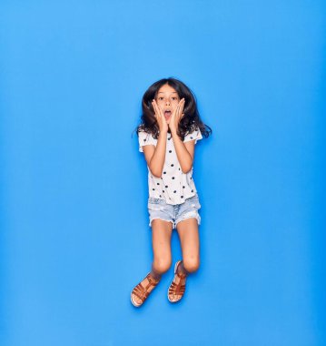 Sevimli İspanyol kız günlük kıyafetler giyip mutlu gülümsüyor. İzole edilmiş mavi arka planda yüzünde gülümsemeyle zıplıyor