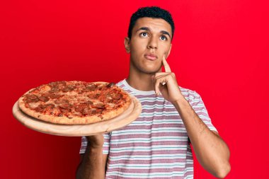 İtalyan pizzası tutan genç Arap adam ciddi bir yüz ifadesiyle eli çenesinde soruyu düşünüyor, kafa karıştırıcı bir fikir hakkında düşünceli. 