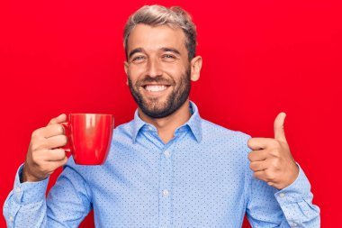 Sakallı yakışıklı sarışın adam izole edilmiş arka planda kırmızı kupa kahve içiyor mutlu ve pozitif gülümsüyor, başparmağıyla mükemmel ve onay işareti yapıyor.