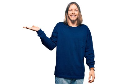 Uzun saçlı yakışıklı beyaz adam sıradan bir kazak giyiyor neşeli bir şekilde gülümsüyor ve avuç içi kameraya bakıyor.. 