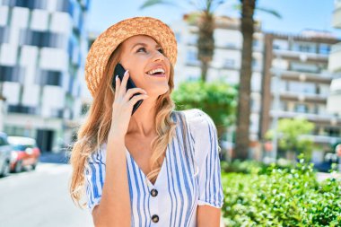 Yaz şapkası takmış, mutlu bir şekilde gülümseyen güzel sarışın bir kadın. Yüzünde gülümsemeyle şehrin caddesindeki akıllı telefondan konuşarak..