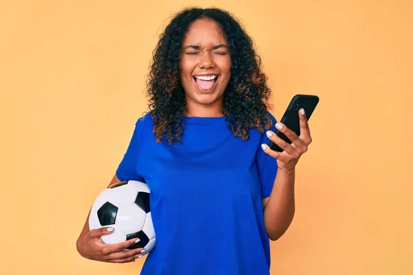 Young african american woman holding football ball looking at smartphone sticking tongue out happy with funny expression.