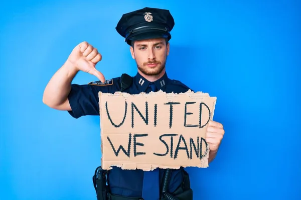 Young caucasian man wearing police uniform holding united we stand banner with angry face, negative sign showing dislike with thumbs down, rejection concept