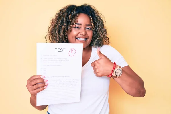 Young african american plus size woman showing a failed exam smiling happy and positive, thumb up doing excellent and approval sign