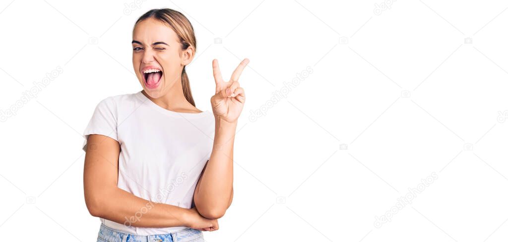 Young beautiful blonde woman wearing casual white tshirt smiling with happy face winking at the camera doing victory sign. number two. 