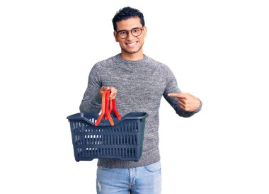 Süpermarket alışveriş sepeti tutan İspanyol yakışıklı genç adam mutlu bir şekilde el ve parmağıyla işaret ediyor. 