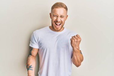 Gündelik beyaz tişört giyen genç beyaz adam kolları havada ve gözleri kapalı bir şekilde başarıyı kutluyor ve şaşırıyor. 