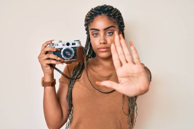 Saç örgülü, elinde antika fotoğraf makinesi olan genç bir Afro-Amerikalı kadın ciddi ve kendinden emin bir ifadeyle dur işareti yapıyor. 